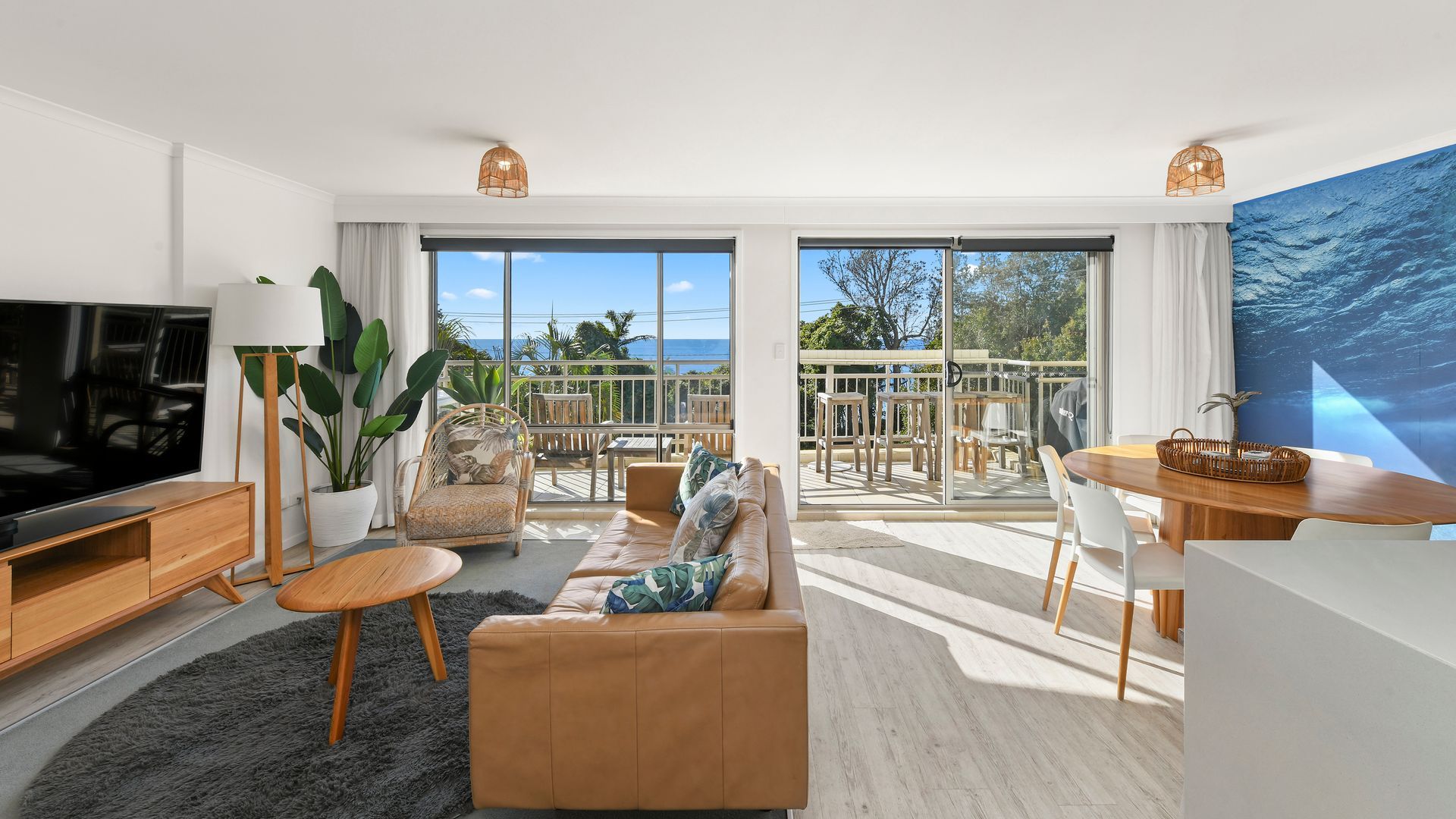 A modern living room with a television, sofa, and dining area, featuring large windows and a view of the outdoors.