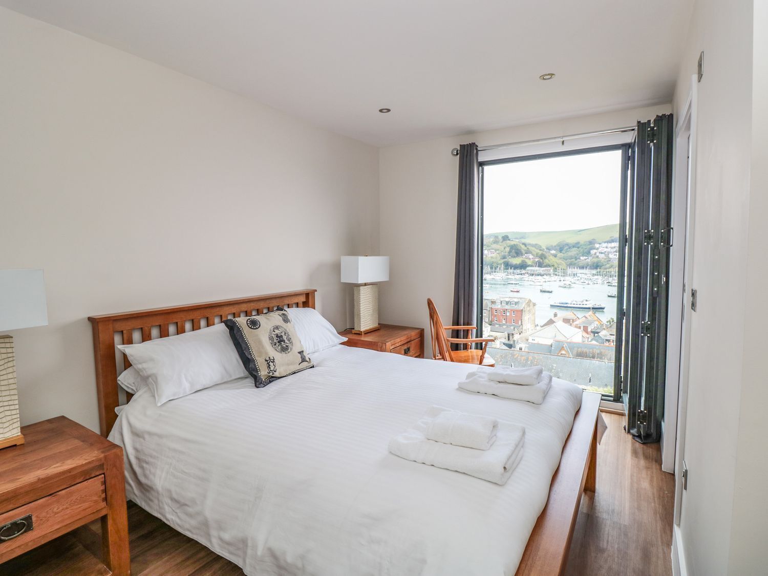 A hotel room with a large bed, a wooden headboard, a chair, a nightstand, a lamp, and a view of a marina through a large window.