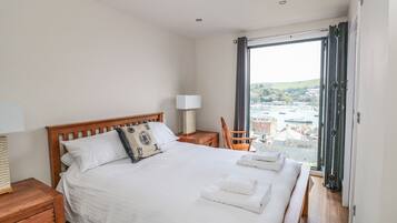 A hotel room with a large bed, a wooden headboard, a chair, a nightstand, a lamp, and a view of a marina through a large window.