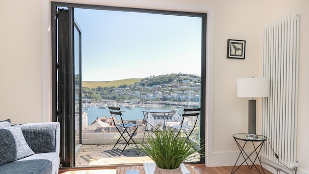 A modern living room with a sofa, a small table with a plant, and a view of a marina and hills. - Ty'r Ddraig (Dartmouth)