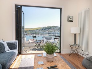 A modern living room with a sofa, a small table with a plant, and a view of a marina and hills. - Ty'r Ddraig (Dartmouth)
