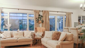 Ein gemütliches Wohnzimmer mit zwei beigefarbenen Sofas, einem kleinen Holztisch und einem Sessel. Große Fenster mit Vorhängen lassen viel natürliches Licht herein und bieten Ausblick auf Grünflächen.