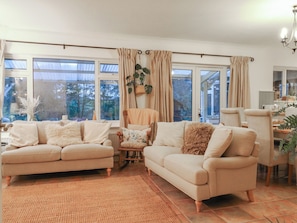 A cozy living room with two beige sofas, a small wooden table, and a chair. Large windows with curtains allow natural light and views of greenery outside. - Berkley Cottage (Colchester)