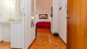 Un couloir étroit doté d’un plancher en bois, d’un canapé rouge, d’un réfrigérateur blanc et d’une porte avec une poignée en laiton.