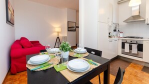 Un coin repas avec une table dressée pour quatre personnes, un canapé rouge, une petite plante en pot et, au fond, une cuisine.