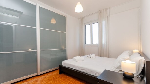 Une chambre d’hôtel moderne dotée d’une grande porte-fenêtre coulissante, d’un lit avec du linge de lit blanc, d’une table de chevet avec une lampe et d’une fenêtre avec des rideaux.