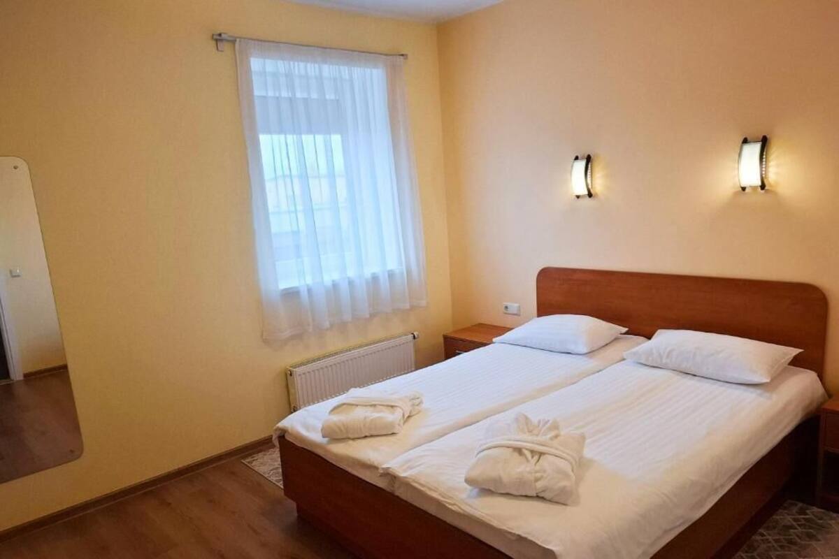 A double bed with white linens and towels, a wooden headboard, two bedside lamps, a window with sheer curtains, and a radiator.