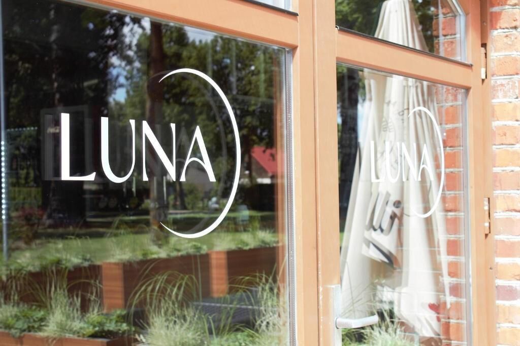 A window with the word "LUNA" in a stylized font, reflecting a garden and a brick wall.
