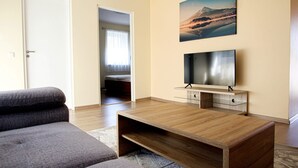 Un salon moderne avec une table basse en bois, un canapé gris, un téléviseur à écran plat posé sur un meuble en bois, et un tableau représentant un paysage de montagne accroché au mur.