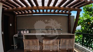 Een terras buiten met een barbecue, twee rieten stoelen en een pergola.