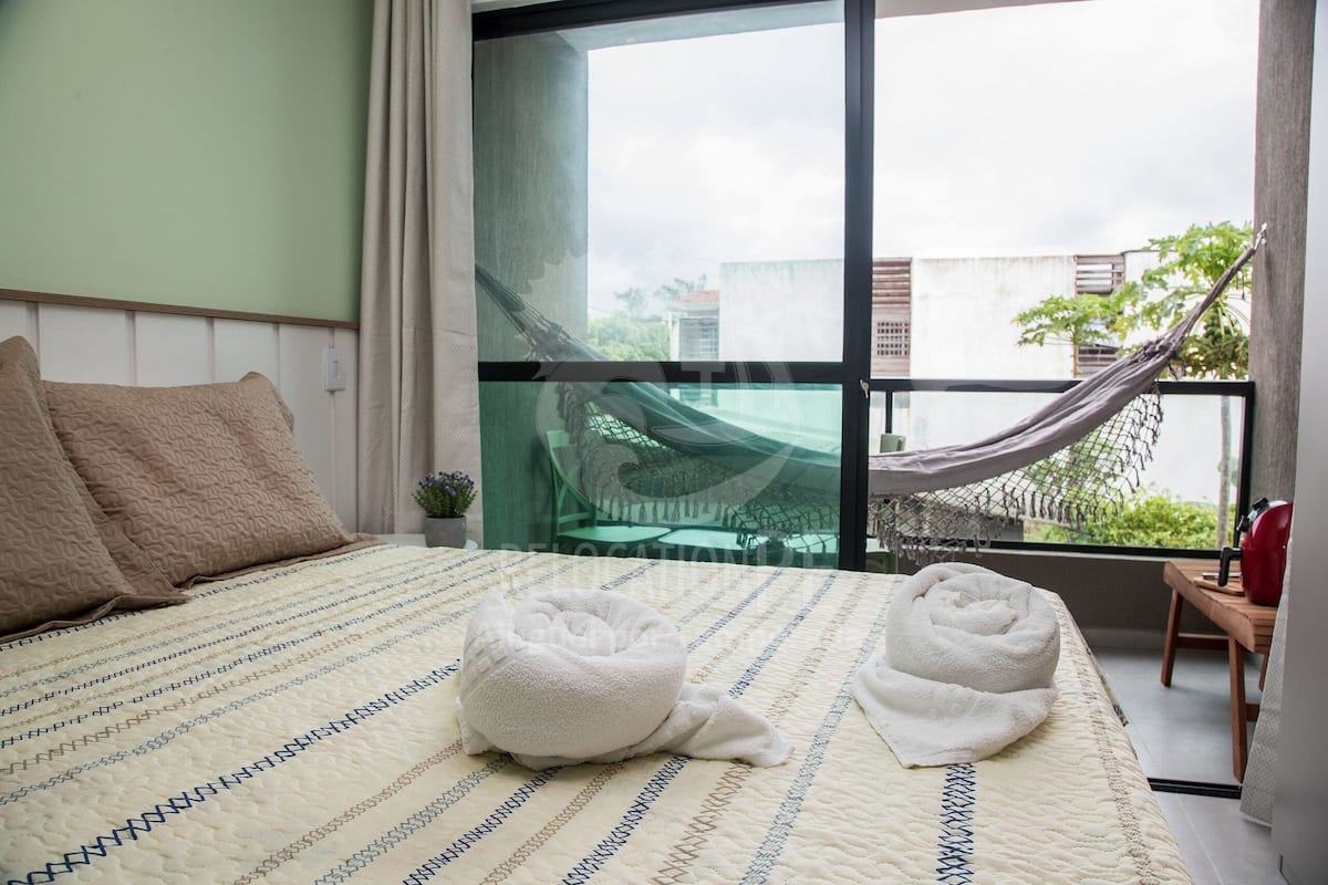 Habitación de hotel con una cama grande, dos toallas, una hamaca y vistas a edificios y vegetación.