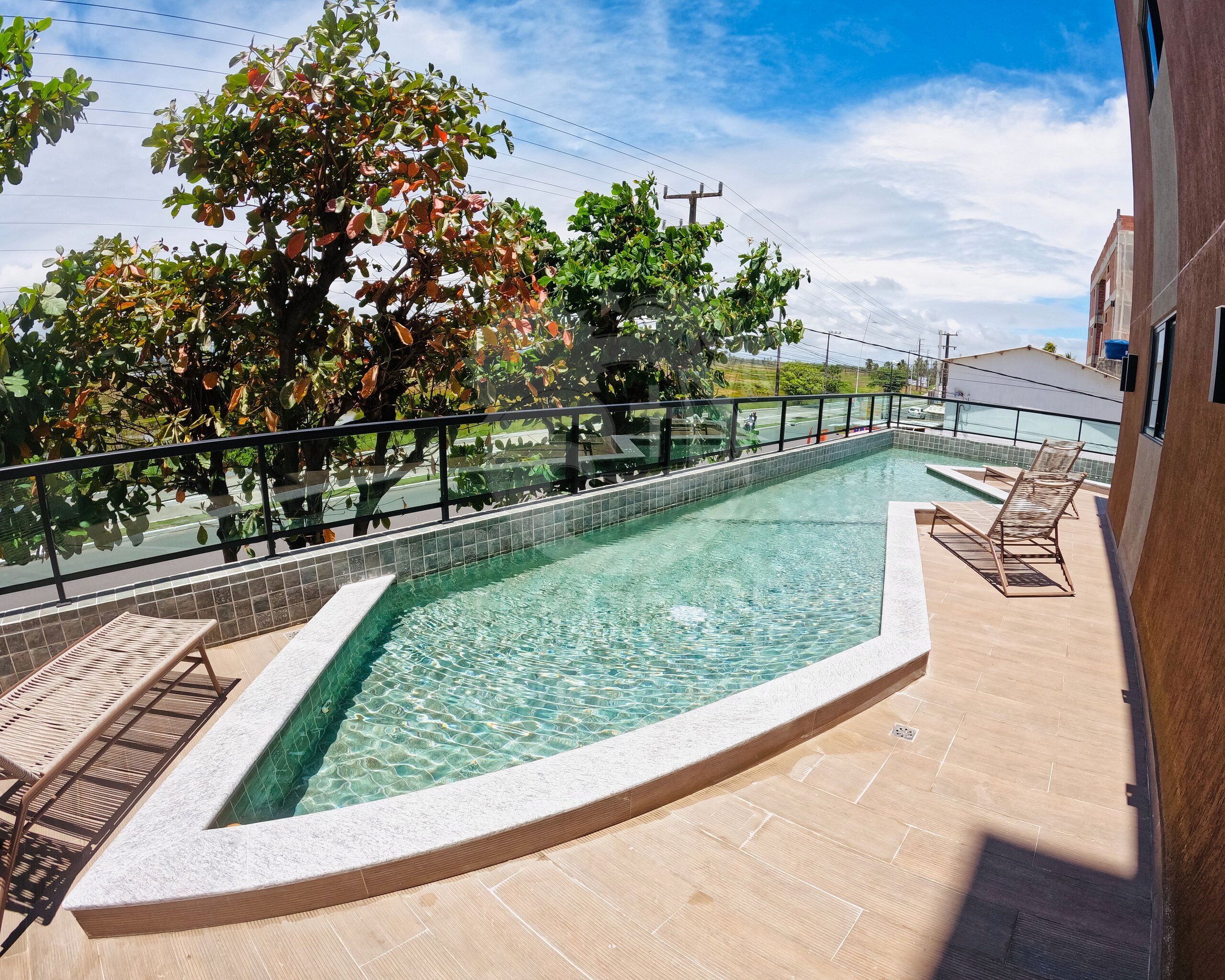A rooftop swimming pool with clear water, surrounded by a tiled deck, and overlooking a road with trees and buildings.