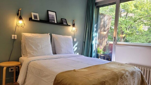 Un dormitorio con cama, mesitas de noche, un ventanal con vistas a árboles e iluminación en la pared.
