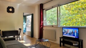 Una sala de estar moderna con un sofá, una mesa de comedor con sillas, un televisor sobre un mueble bajo y ventanas amplias con vistas a árboles.