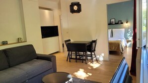 Una sala moderna con un sofá, una mesa de comedor con sillas, una mesita redonda y un televisor. Se ve una cama al fondo con un ventanal.