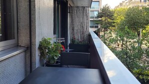 Un balcón con una mesa y sillas negras, una planta en maceta y vistas a una zona residencial con árboles y edificios.