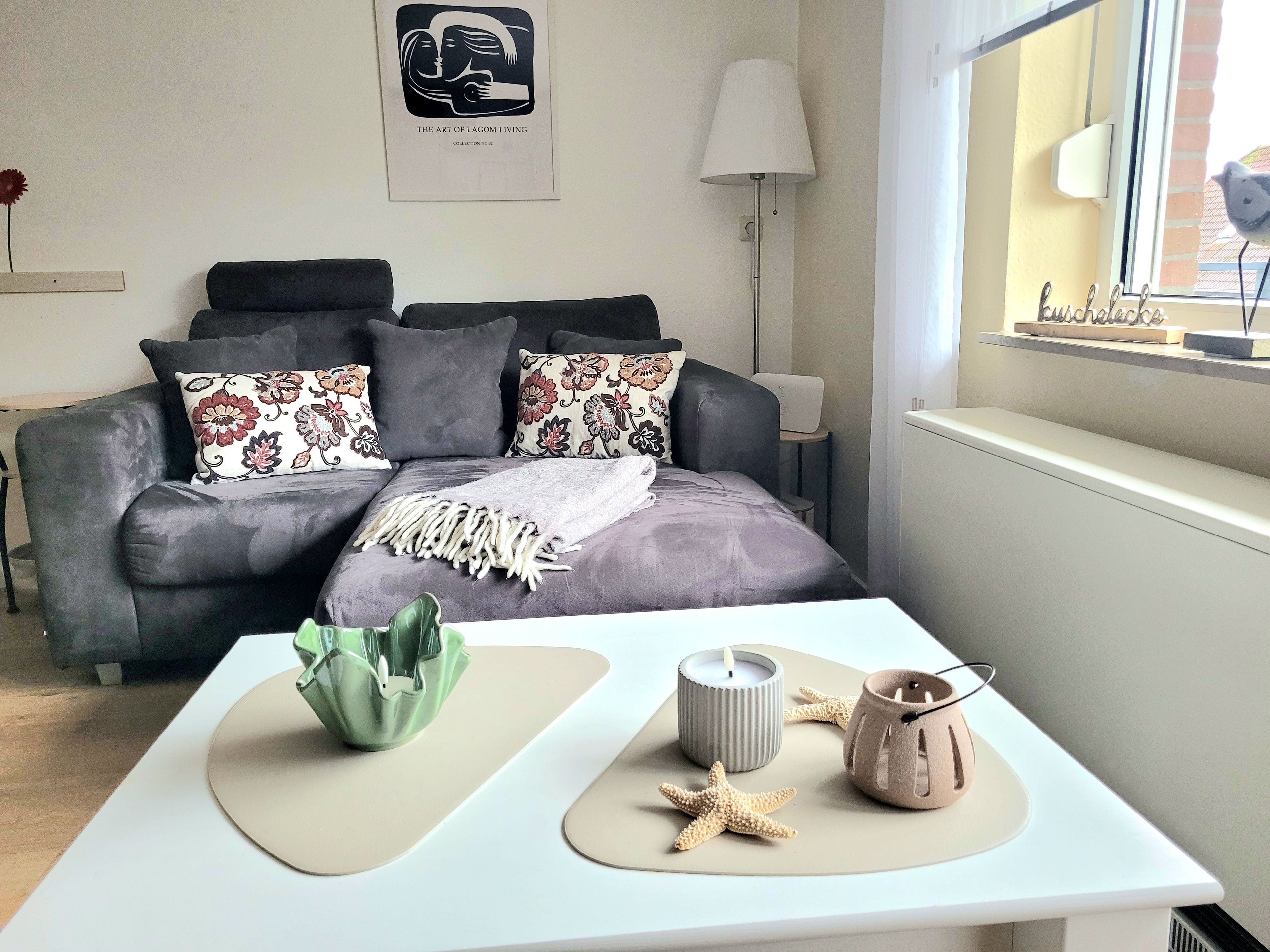 Uma sala de estar moderna com um sofá cinzento, almofadas florais, uma mesa de centro branca com objetos decorativos e uma obra de arte emoldurada na parede.
