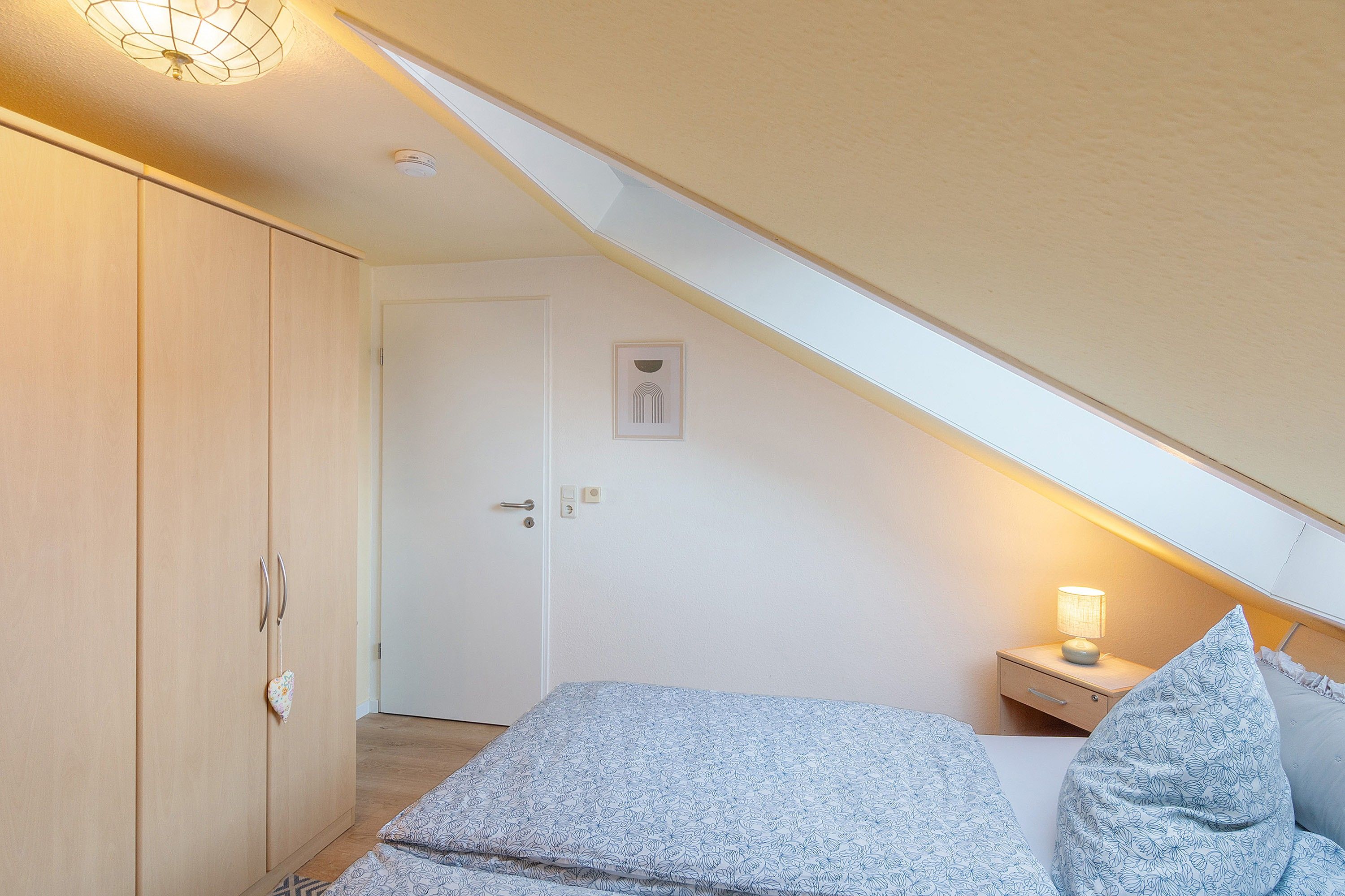 A bedroom with a sloped ceiling, a bed with patterned bedding, a bedside table with a lamp, and a wardrobe.