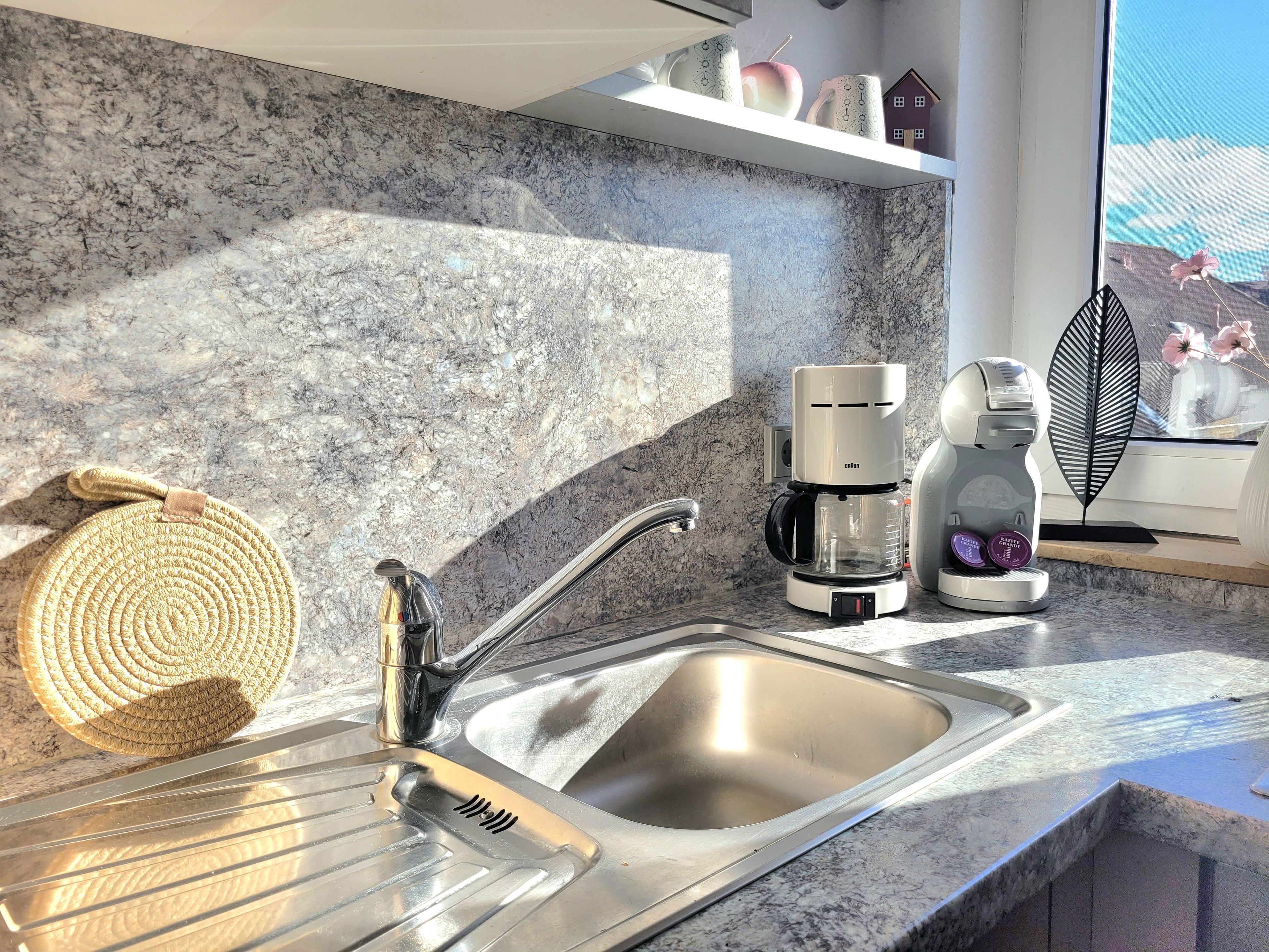 A kitchen with a stainless steel sink, a coffee maker, and a decorative leaf.