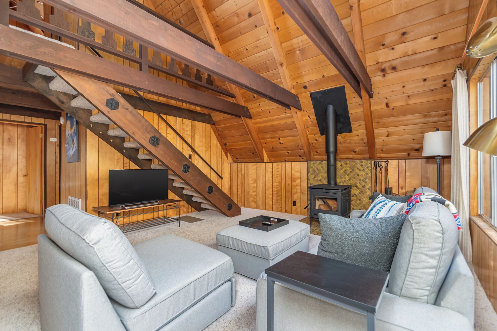Acogedor interior de cabaña de madera con escalera, chimenea, televisor y asientos cómodos.