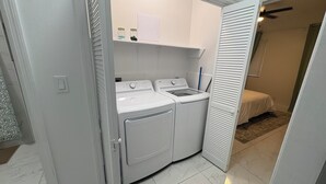 Una lavanderia con lavatrice e asciugatrice, un piccolo ripiano con barattoli, e un letto nella stanza confinante.