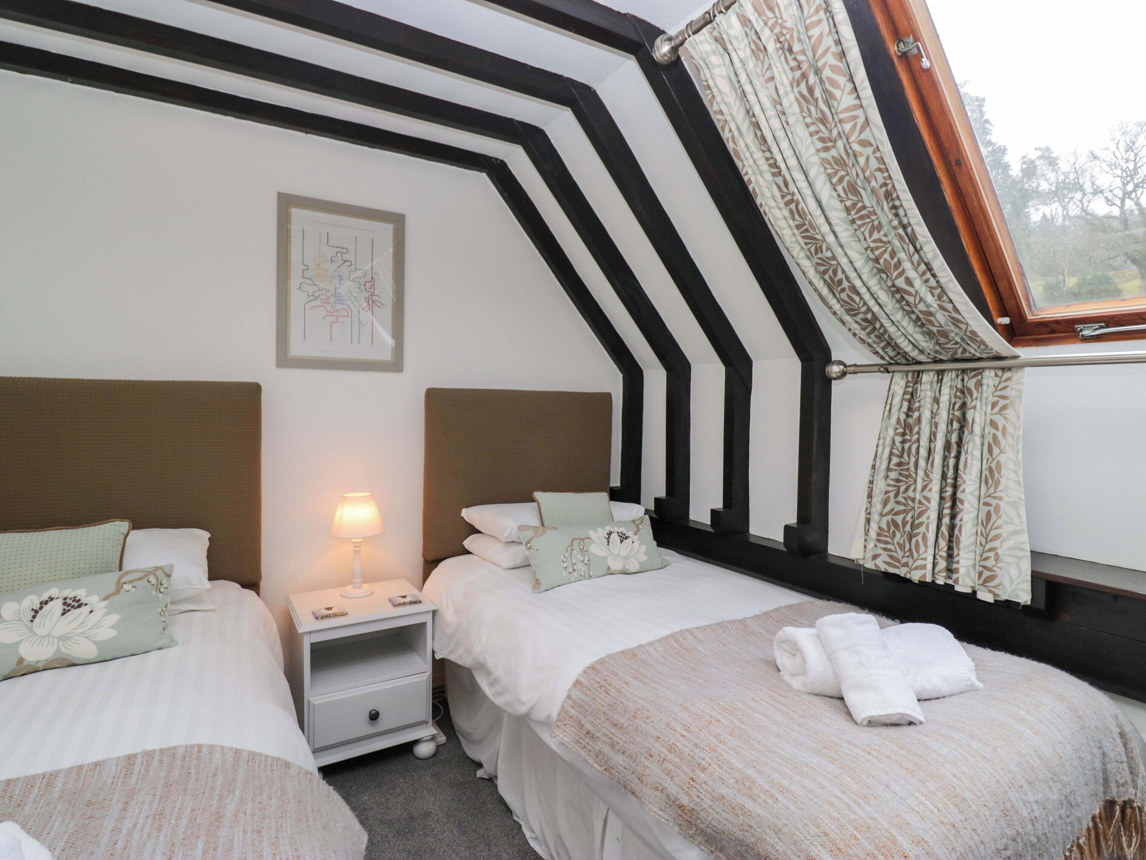 Une chambre avec deux lits, une table de chevet, une lampe, un tableau encadré et une fenêtre avec des rideaux.