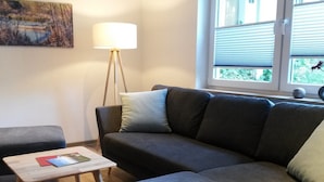 A living room with a sofa, a small table, a floor lamp, and a window with blinds. - Schrichten Apartment on the Edge of the Forest (Schmallenberg)