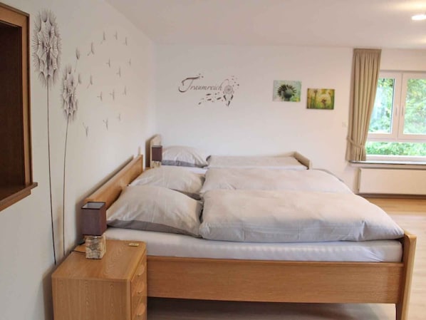 A bedroom with a wooden bed, a nightstand, a wall-mounted lamp, and a window with curtains.