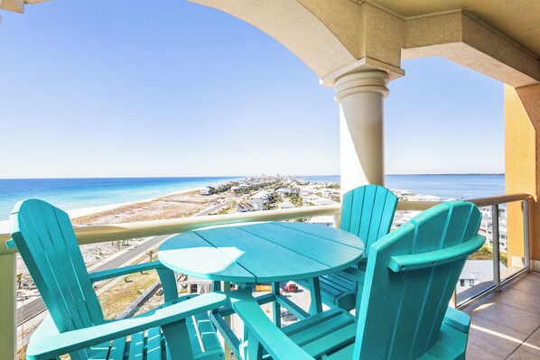 Ein Balkon mit türkisfarbenen Stühlen und einem Tisch mit Blick auf einen Strand und den weiten Ozean sowie entfernte Gebäude.