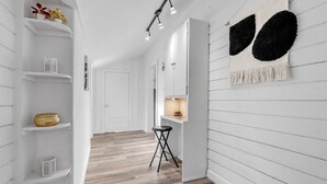 A hallway with white walls, wooden flooring, and a black stool. Shelves with decorative items and a hanging tapestry are visible. - Charming Downtown Buda Cottage | Walk to Dining, Shops & Nightlife (Buda)