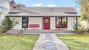 A single-story house with a red door, a front porch with a red bench, and a number '220' on the front steps. - Charming Downtown Buda Cottage | Walk to Dining, Shops & Nightlife (Buda)