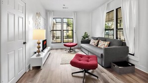 A modern living room with a grey sofa, red ottoman, and a white side table with a lamp. - Charming Downtown Buda Cottage | Walk to Dining, Shops & Nightlife (Buda)