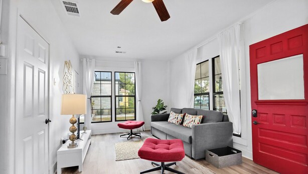 A modern living room with a grey sofa, red ottoman, and a red door. - Charming Downtown Buda Cottage | Walk to Dining, Shops & Nightlife (Buda)