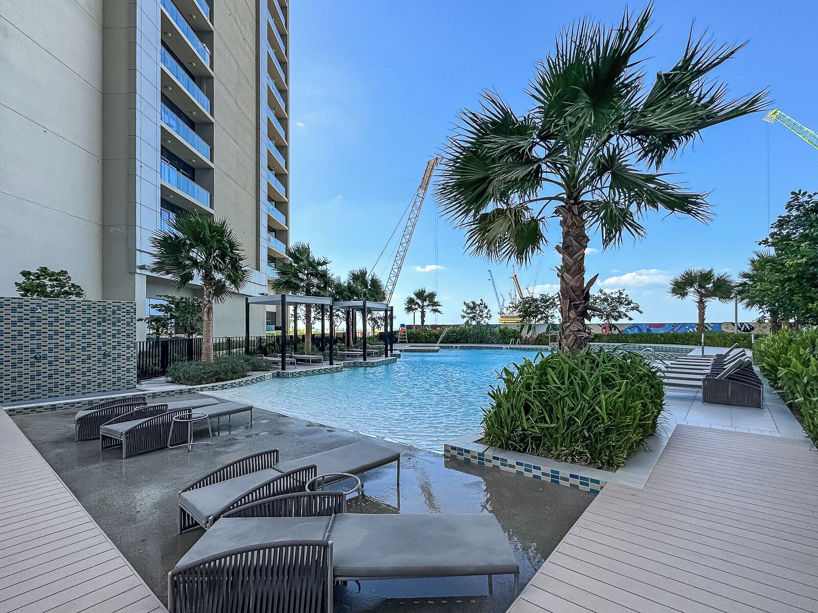 A pool area with lounge chairs and a palm tree, surrounded by a high-rise building and construction cranes in the background.
