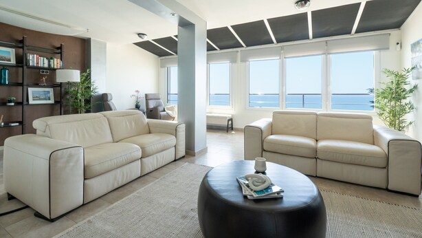 Un soggiorno moderno con due divani beige, un tavolino da caffè nero e una libreria. Ampie finestre offrono vista sul mare.
