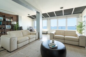 Un salon moderne avec deux canapés beige, une table basse noire et une bibliothèque. De grandes fenêtres offrent une vue sur la mer.