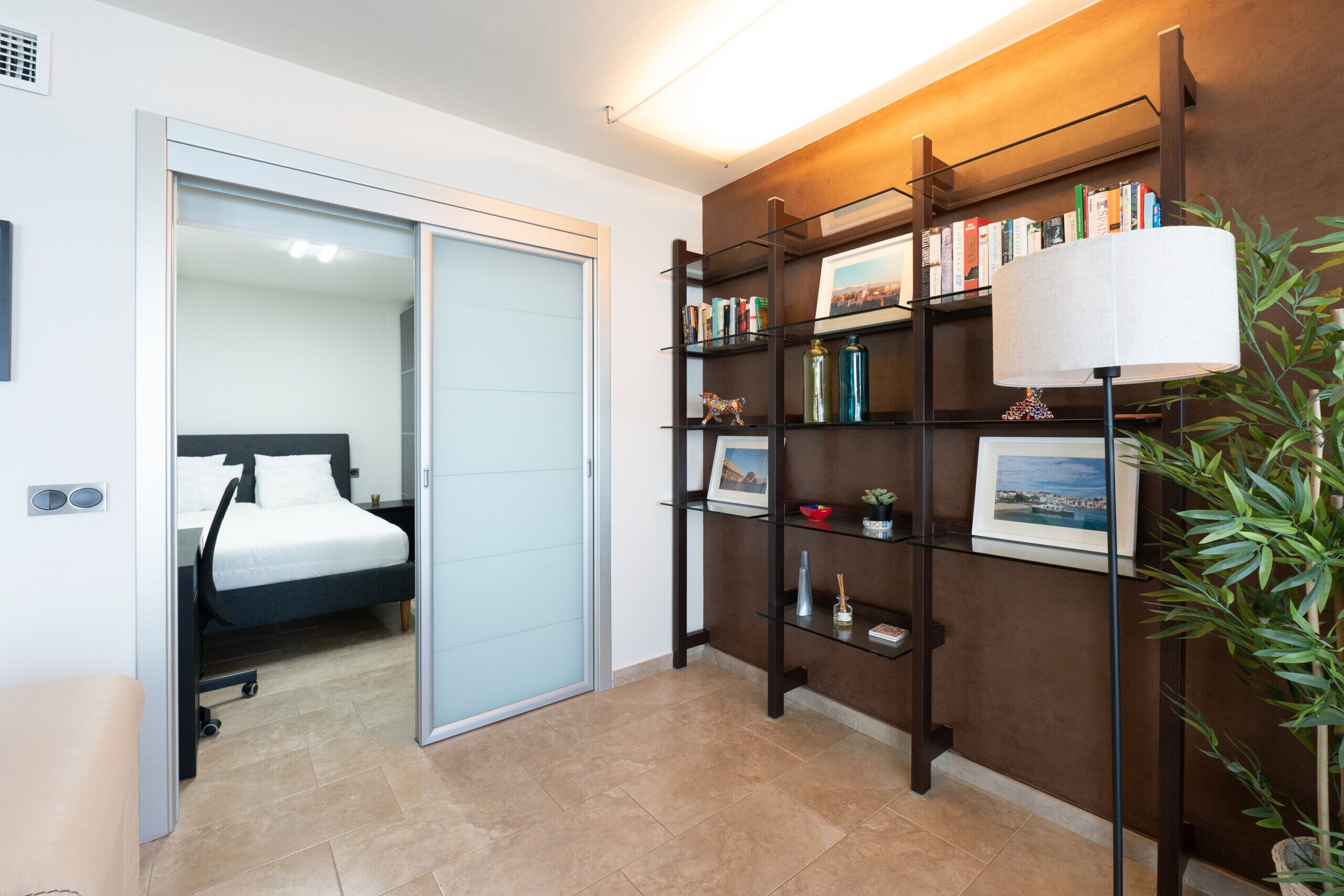 Habitación de hotel con cama, puerta corrediza, estantería con libros y objetos decorativos, lámpara y planta en maceta.
