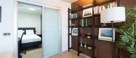 Habitación de hotel con cama, puerta corrediza, estantería con libros y objetos decorativos, lámpara y planta en maceta.