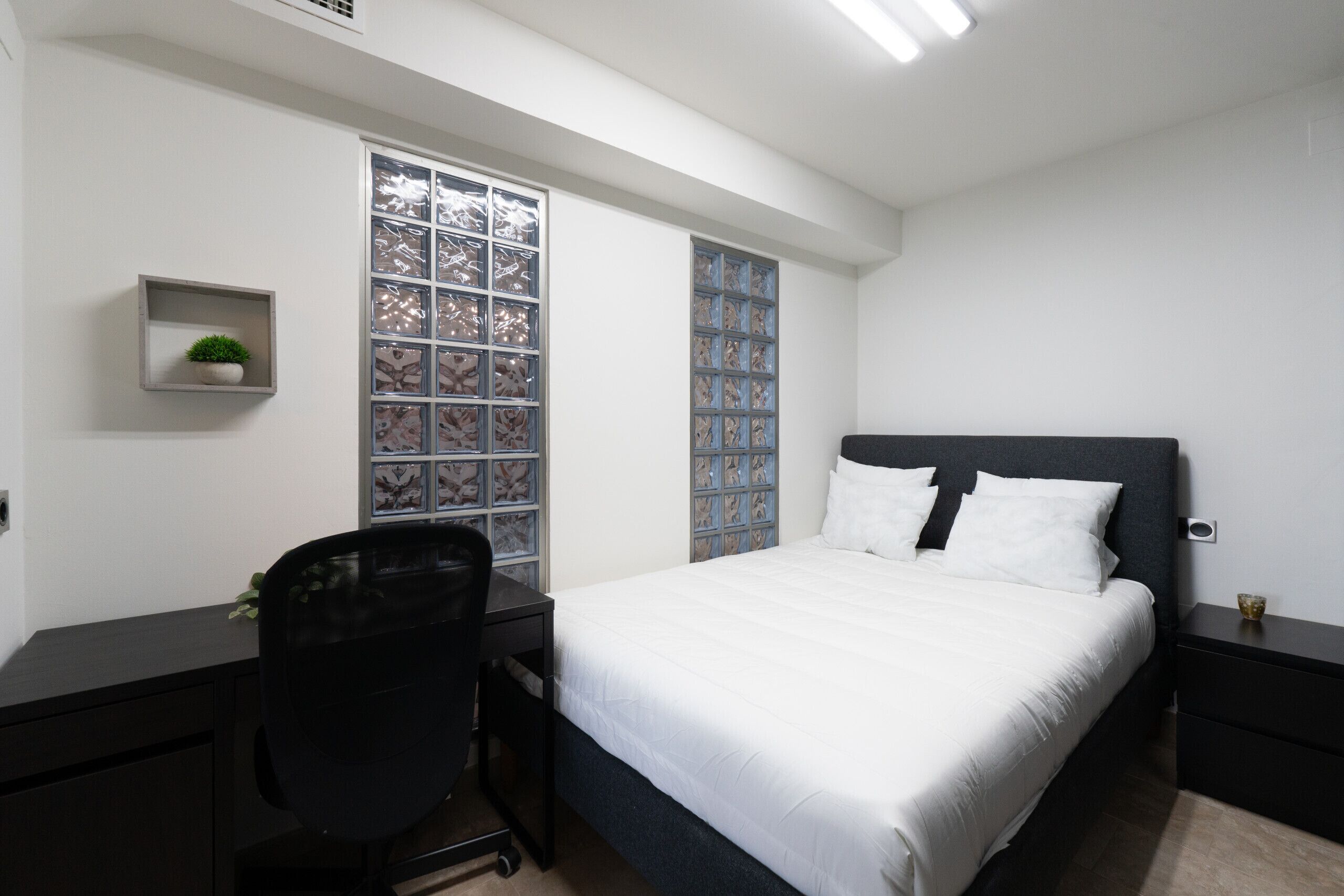 A bedroom with a bed, a desk, a chair, a small shelf with a plant, and a window with patterned glass.