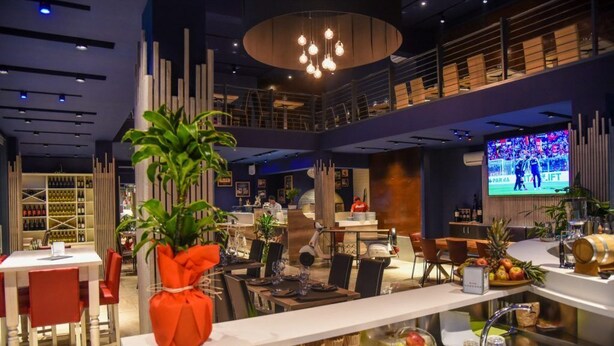 A modern restaurant with a high ceiling, a central plant, and a TV screen displaying a sports event. - HOTEL BUSINESS (Casoria)