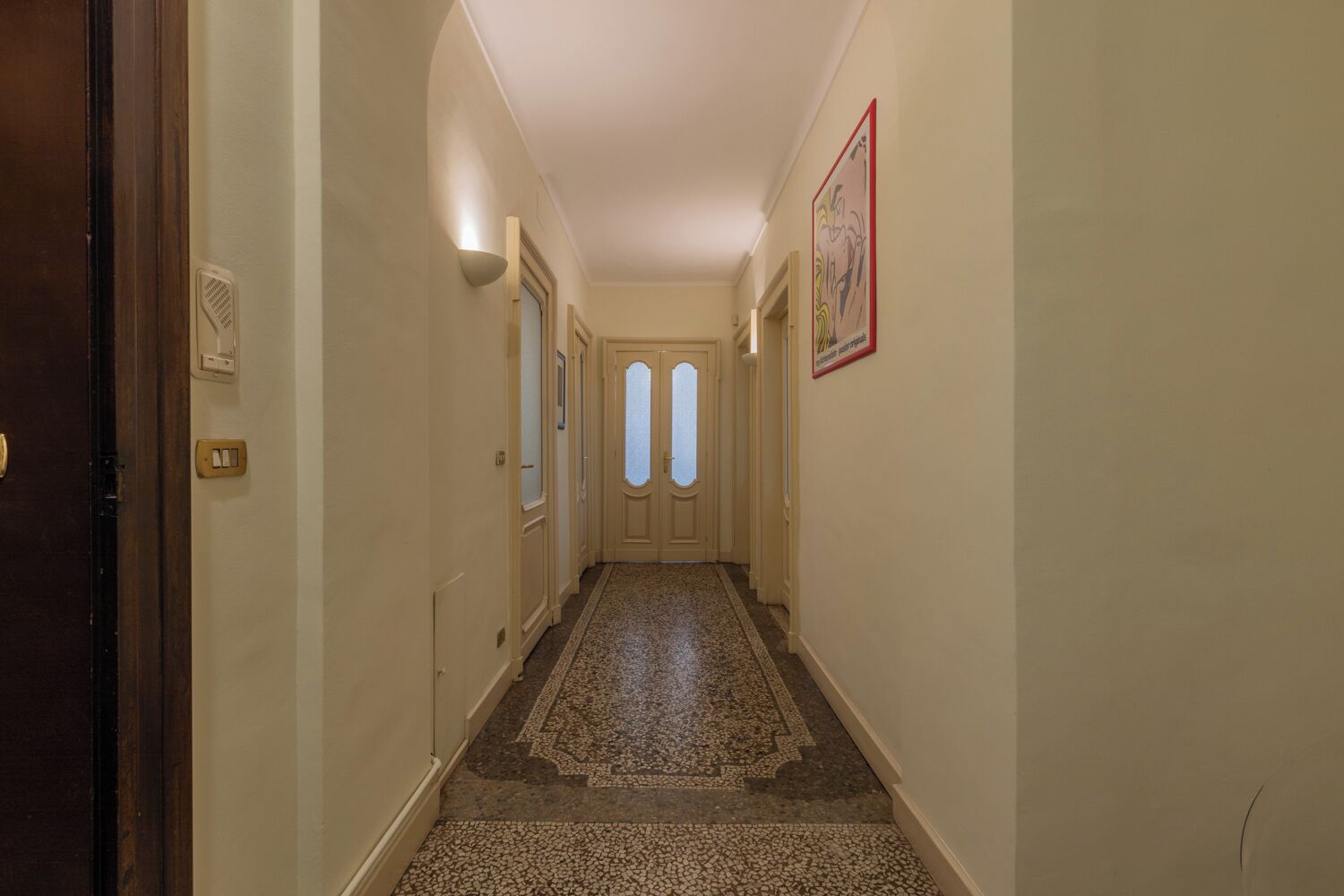 A hallway with a patterned carpet, a door at the end, and a wall-mounted light fixture.