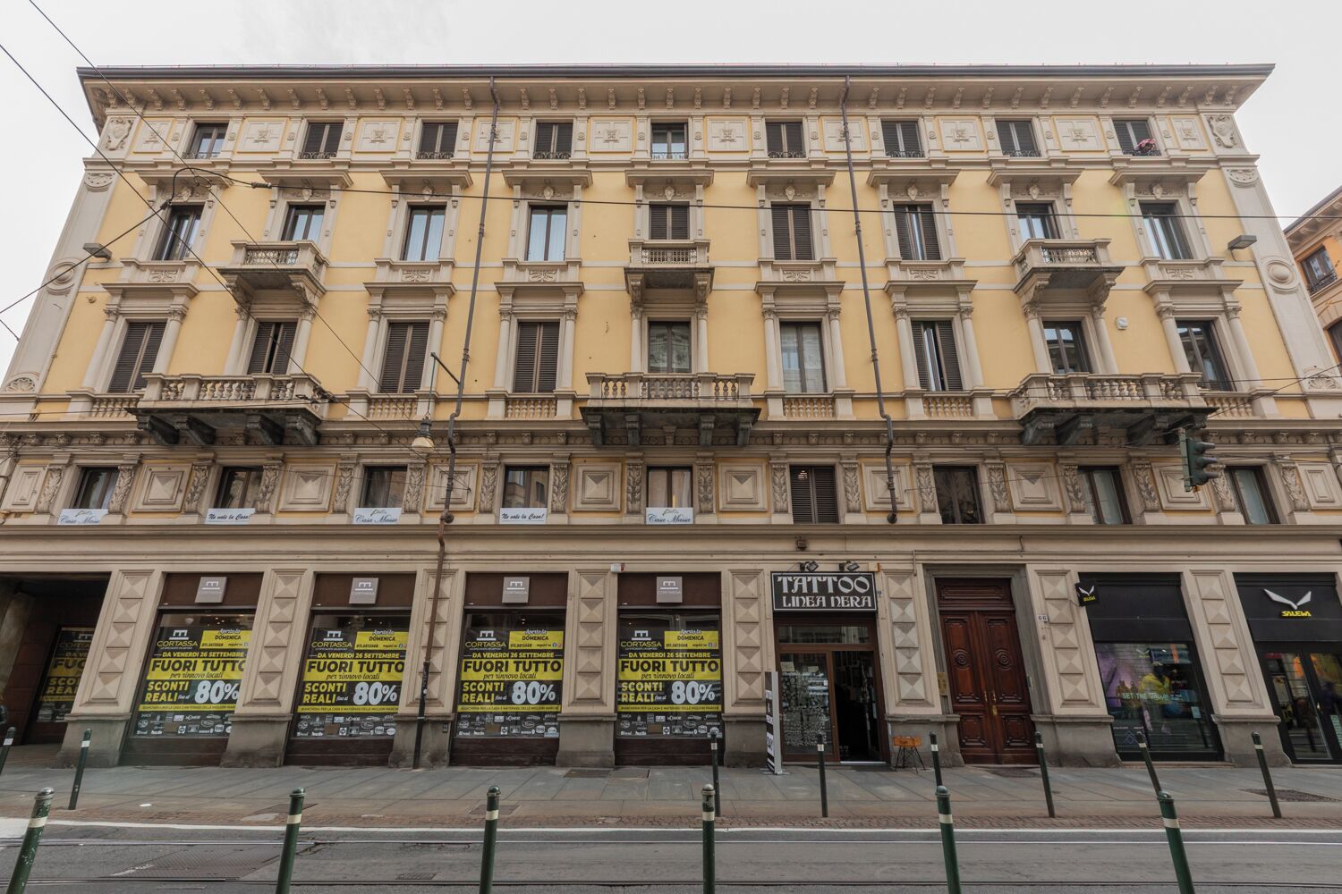 A multi-story building with a yellow facade, multiple balconies, and storefronts including a tattoo parlor.