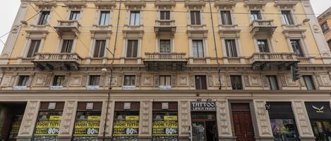 A multi-story building with a yellow facade, multiple balconies, and storefronts including a tattoo parlor.