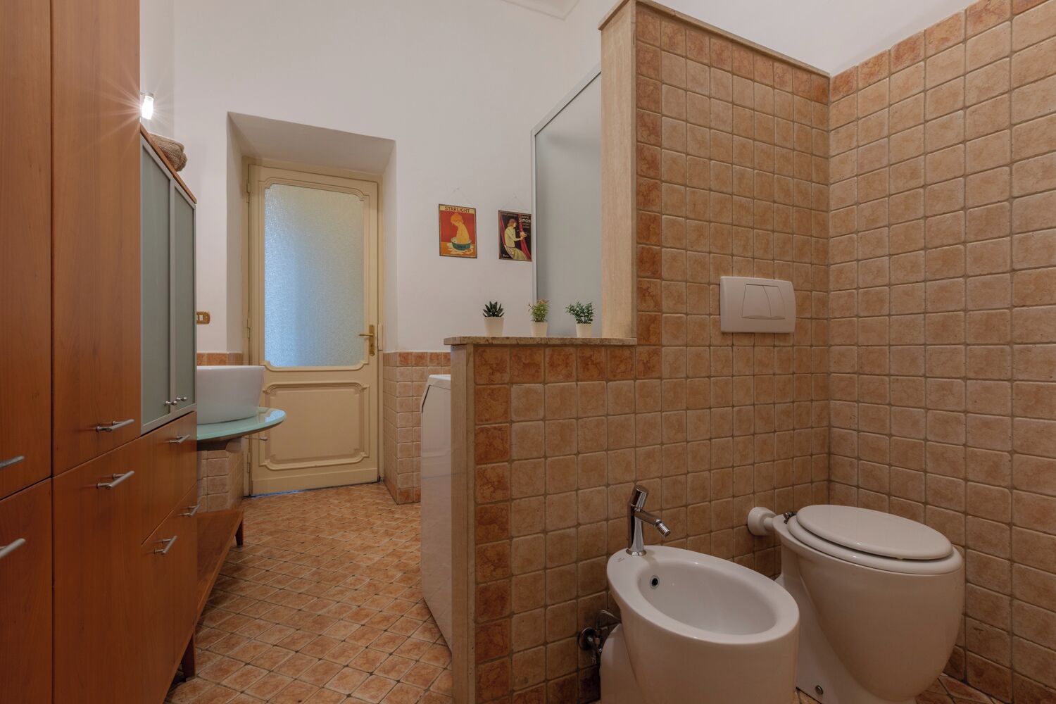 Une salle de bain comprenant des toilettes, un bidet et un lavabo. On y trouve un meuble en bois avec des tiroirs et un miroir. Les murs sont recouverts de carreaux dans un motif marron.