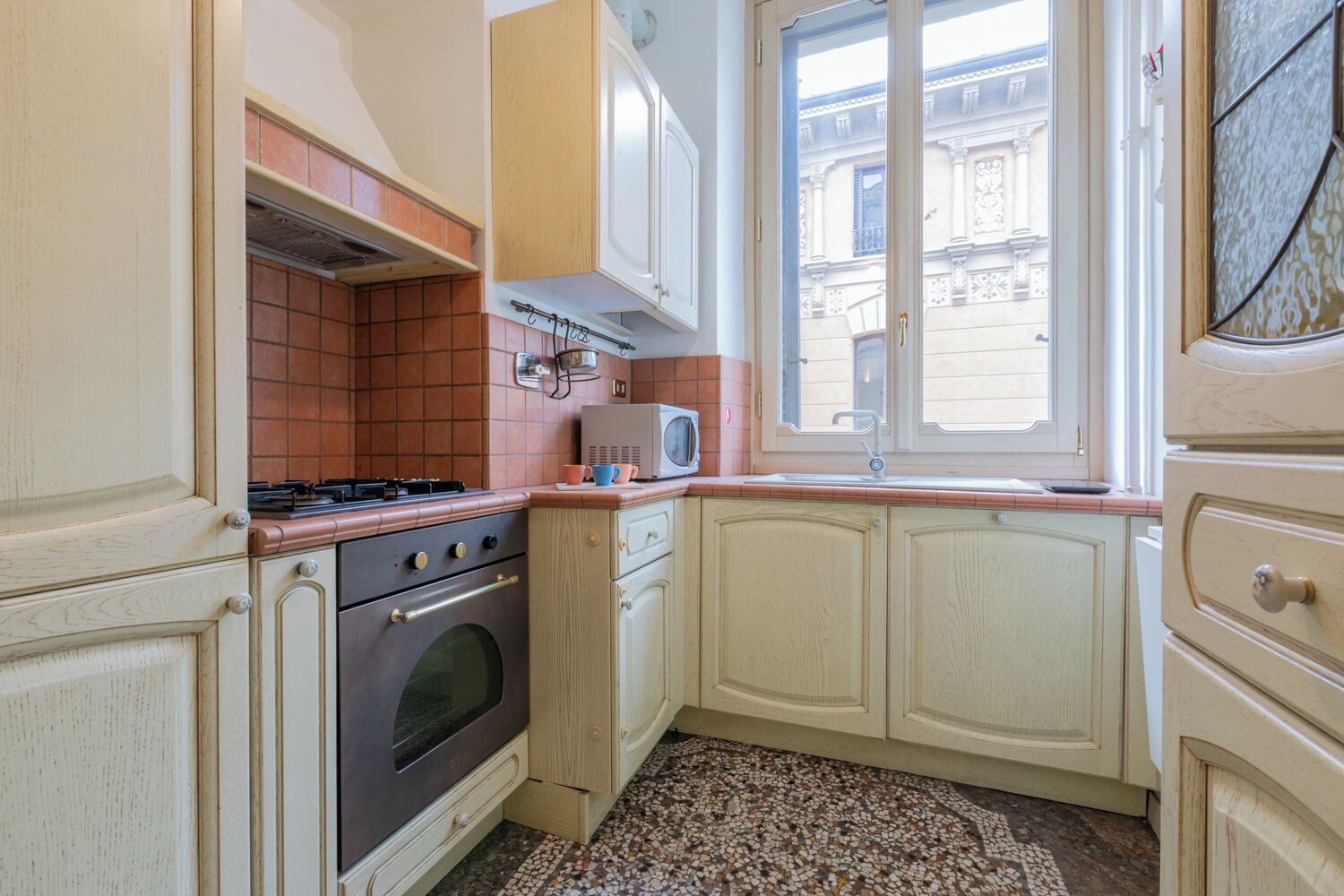 A compact kitchen with a gas stove, oven, and a microwave on the countertop. There is a window providing natural light and a view of the building outside.