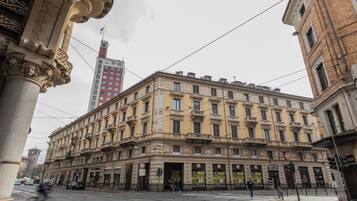 Un edificio histórico con detalles arquitectónicos ornamentales, varias tiendas a pie de calle y una torre destacada al fondo.