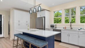 Een moderne keuken met witte kasten, een centraal eiland met barkrukken, roestvrijstalen apparatuur en grote ramen met uitzicht op groen.