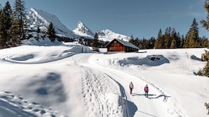 Une cabane en bois dont le toit est recouvert de neige, entourée de montagnes et d’arbres également recouverts de neige.