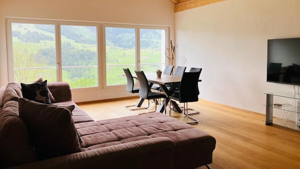 Un salon spacieux doté d’une grande fenêtre, d’un plafond en bois, d’un canapé marron, d’une table à manger avec des chaises et d’un téléviseur à écran plat.