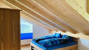 Une chambre à coucher aménagée dans un espace de type « loft », avec un plafond en bois, un lit avec une literie bleue et une fenêtre donnant sur un paysage verdoyant.